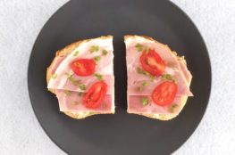 Open ham sandwich on white bread with tomato and chive garnish on grey plate and white background