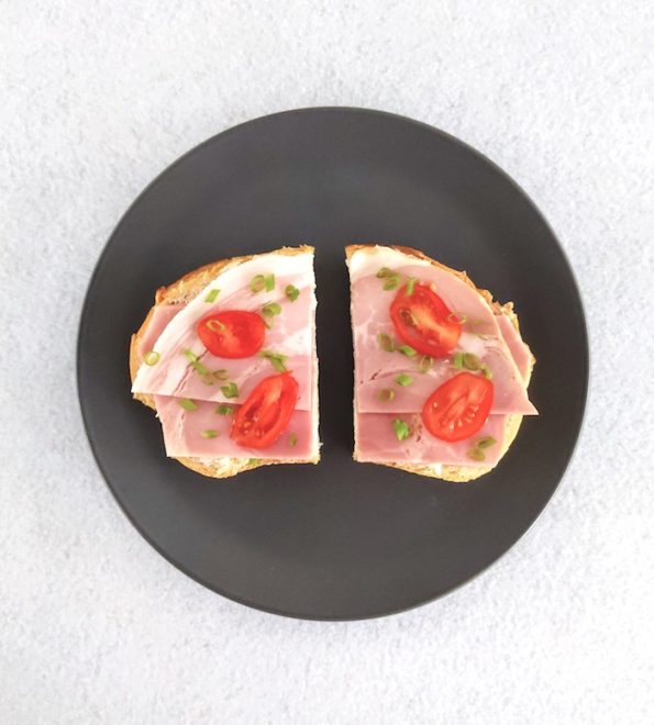 Open ham sandwich on white bread with tomato and chive garnish on grey plate and white background