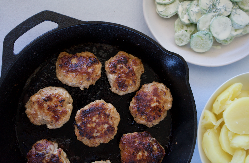 Latvian meatballs on a black cast iron pan with bowls of boiled potato and cucumber salad